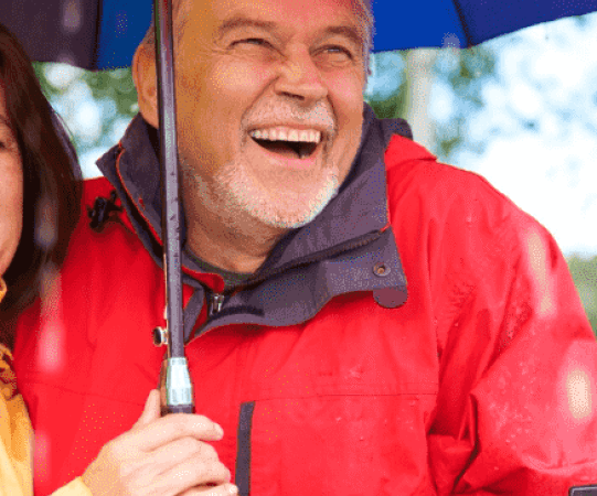 Smiling older couple under umbrella in rain, showcasing happiness and support, relevant to Canadian immigration services.