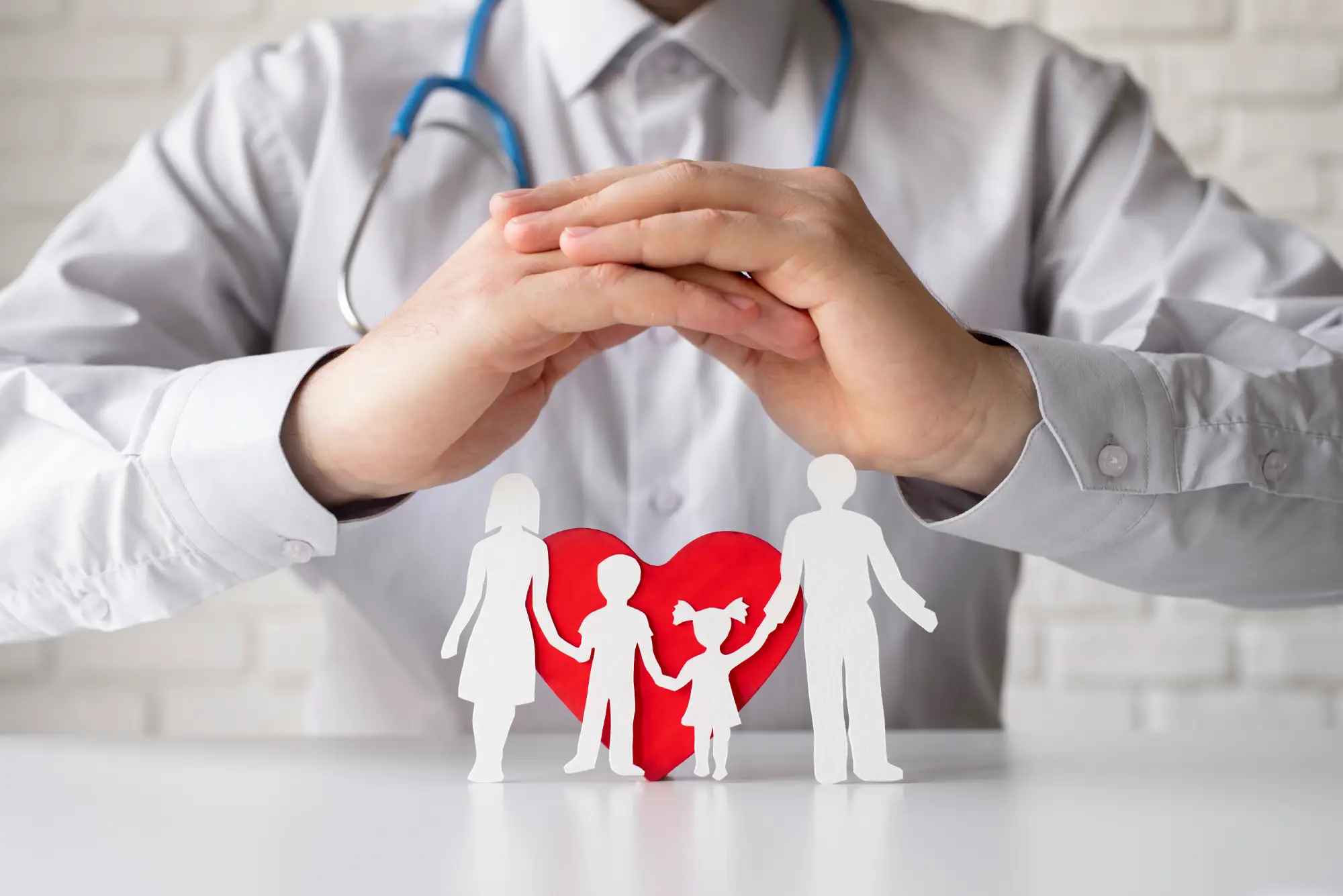 Close-up of a doctor with a stethoscope, hands forming a protective gesture over paper cutouts of a family in front of a red heart, symbolizing healthcare and social services protection.